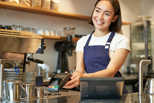 Smiling asian girl barista, giving client POS terminal, help pay with credit card, standing at counter in coffee shop, working in cafe
