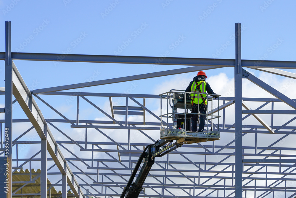 Worker in self propelled boom lift basket in uniform on a building steel framework background - obrazy, fototapety, plakaty 