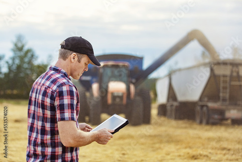 A farmer using his tablet to help manage the wheat harvest while a combine is offloading a full load of grain to a grain buggy: Alcomdale, Alberta, Canada photo