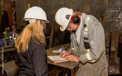 A female plant manager provides administrative documents for signing by an employee for job tracking in a fabrication plant; Innisfail, Alberta, Canada
