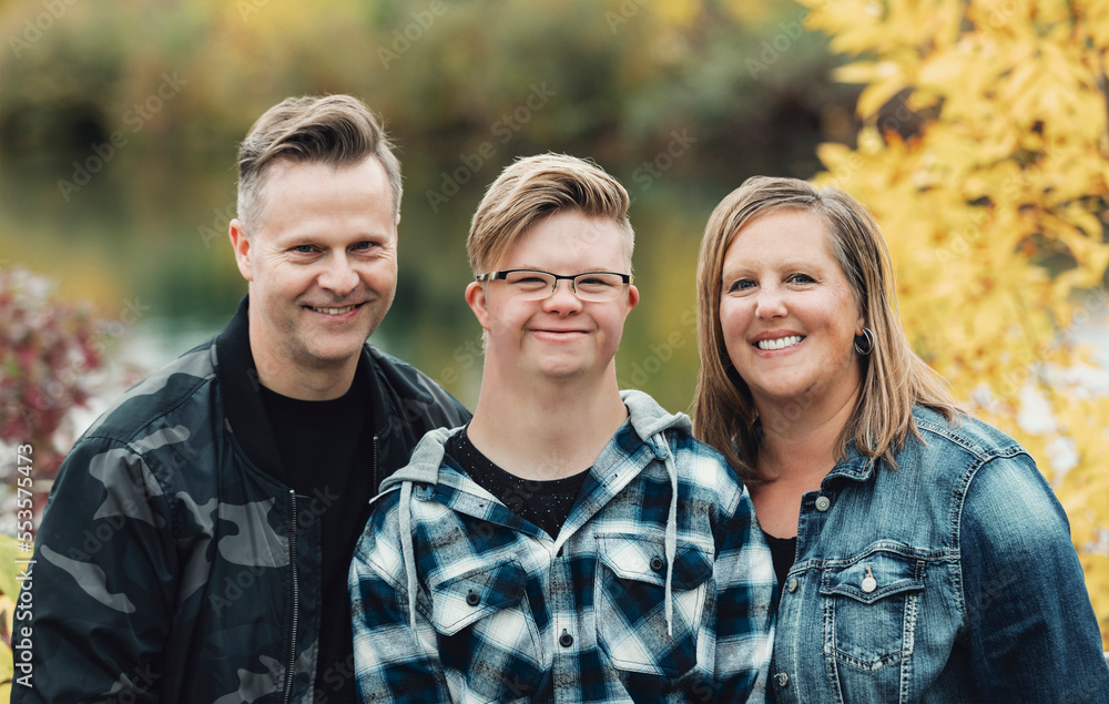 A young man with Down Syndrome posing for a family portrait with his father and mother while enjoying each other's company in a city park on a warm fall evening; Edmonton, Alberta, Canada A young man with Down Syndrome posing for a family portrait with his father and mother while enjoying each other's company in a city park on a warm fall evening; Edmonton, Alberta, Canada