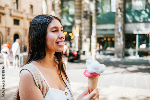 Happy Hispanic female carrying ice cream and looking away with smile on blurred background of street on sunny summer day in Barcelona, Spain