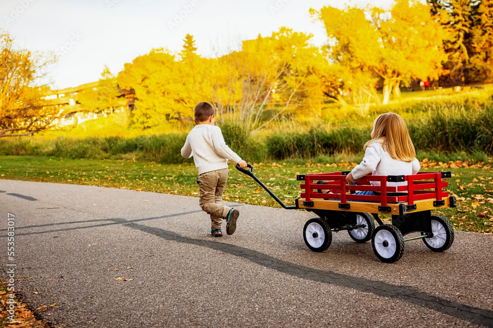 Young boy pulling his sister in a wagon at a city park along a river ...
