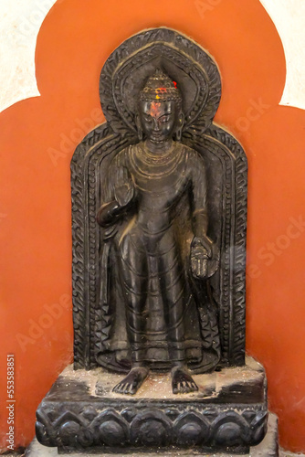 Small black metal Buddha statue stands by red wall. Traces of paints remained on the head after the offerings. Culture of India theme.