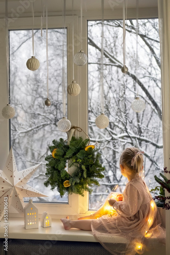 A little girl in a dress sits on the windowsill and looks at the snow. Сhristmas snowy day