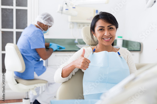 Portrait of satisfied Latina in dental office showing approval gesture with thumbs up after dental treatment