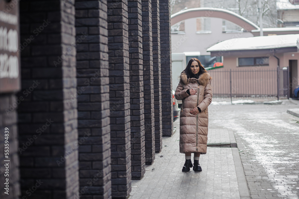 Beautiful young pregnant woman in a brown coat, in the background a brick wall