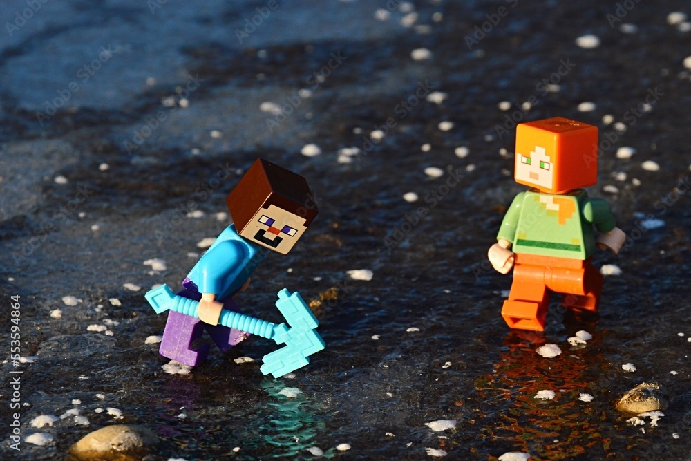Obraz premium LEGO Minecraft figures of Steve and Alex on thick layer of real ice Steve is trying to break ice with his diamond pickaxe.