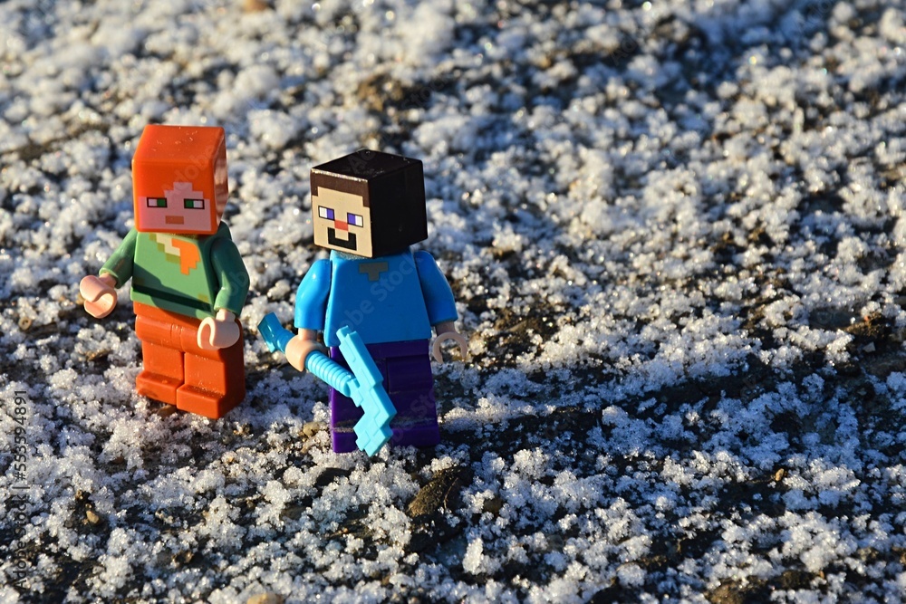 Naklejka premium LEGO Minecraft figures of Steve and Alex on a walk on dirt road covered with first flakes of snow, morning sunshine. Steve is holding diamond pickaxe