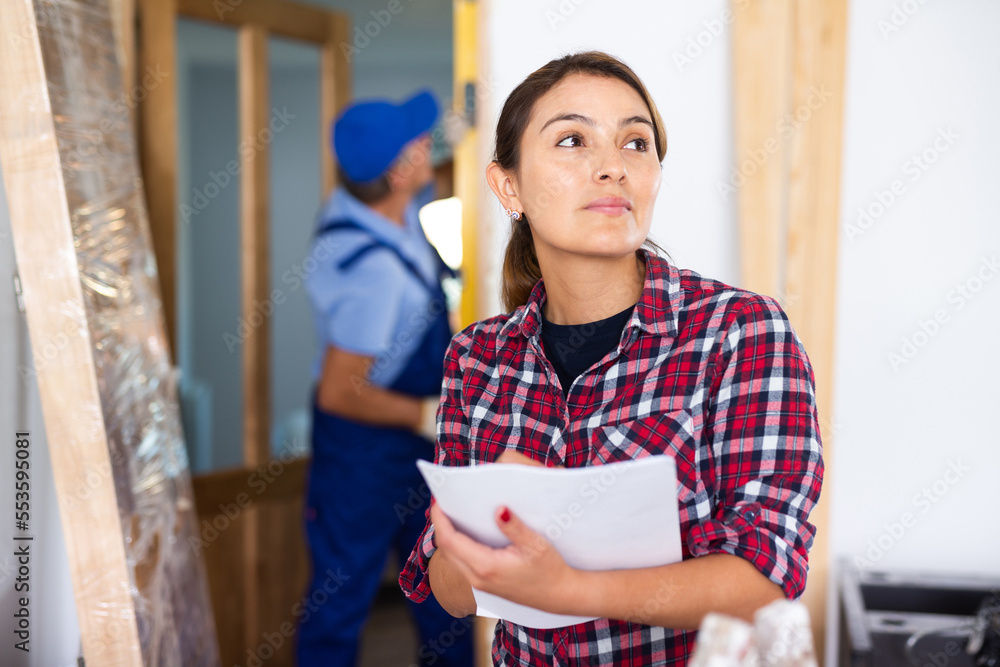 Fototapeta premium Latin woman standing in building site in apartment and inspecting rooms, checking project documentation.