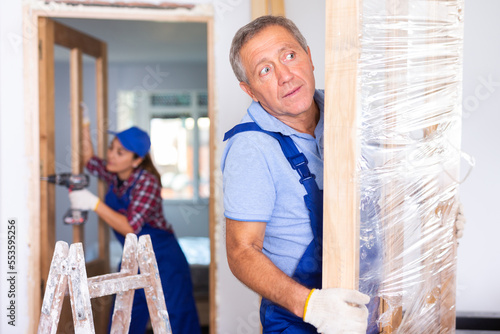 Professional male craftsman in overalls dragging the door construction into an apartment