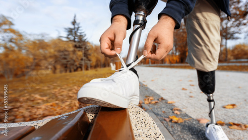 View of a man with prosthetic legs and white sneakers. Tying shoelaces with his foot on the bench in a park. Fallen yellow leaves around