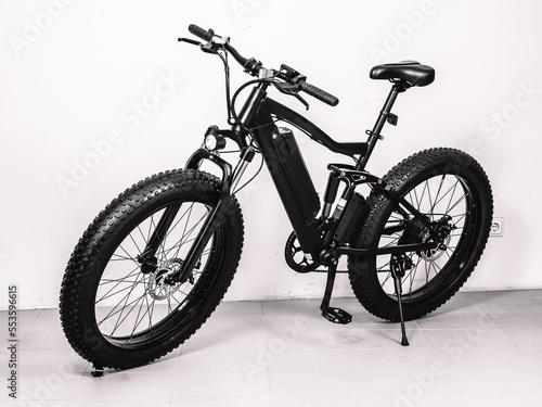Modern black bicycle with thick wheels and an electric motor. White wall.