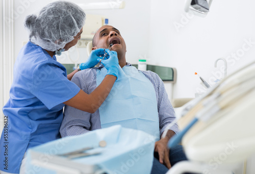 Hispanic man sitting in dental chair, getting anesthetic injection before teeth treatment by professional female dentist