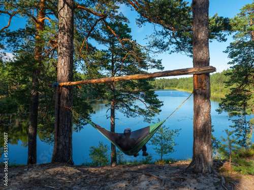 A man rests in a hammock and enjoys a view of the river and the forest. Summer nature.