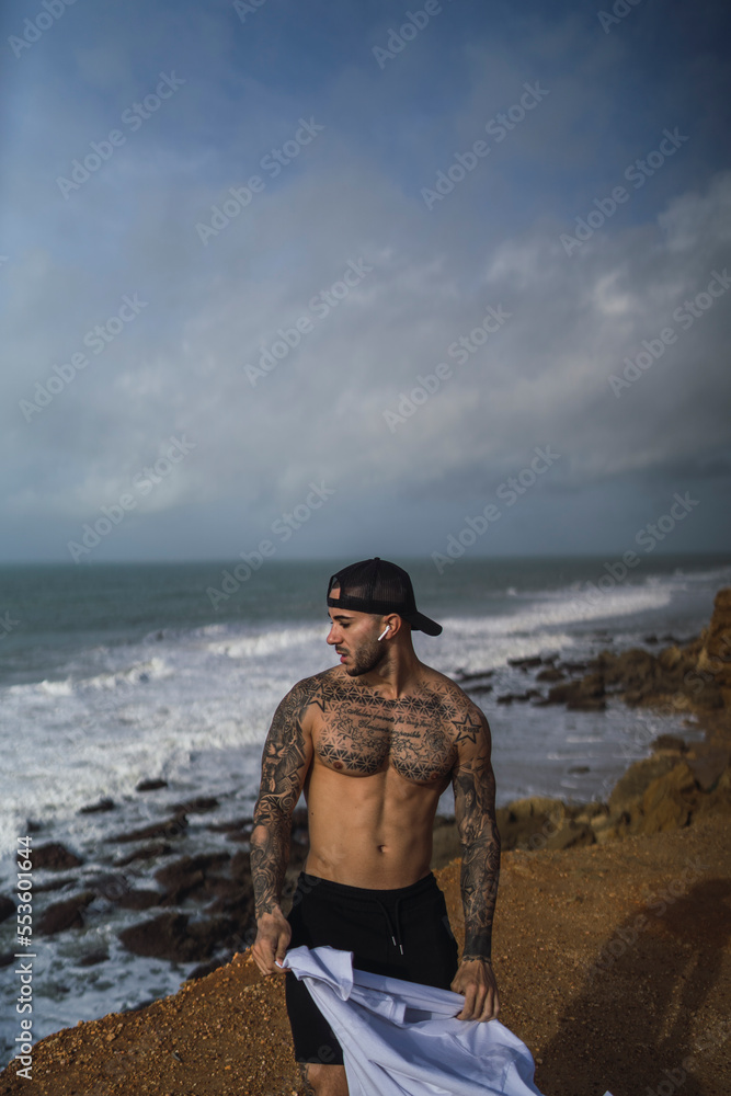 Chico joven musculado y tatuado posando en acantilado de una playa sin camiseta