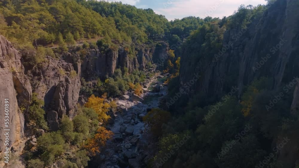 Flight over the shallow river bed between green rocks. Mountain landscape. Blue sky and clouds. Aerial drone footage