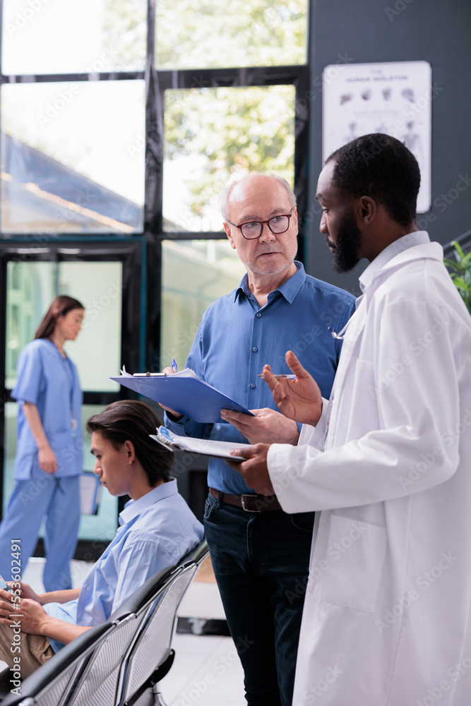 African american doctor discussing medical expertise to senior patient during checkup visit consultation in hospital reception. Medic explaining healthcare treatment while man signing insurance papers African american doctor discussing medical expertise to senior patient during checkup visit consultation in hospital reception. Medic explaining healthcare treatment while man signing insurance papers