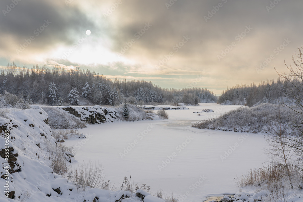 Estonian winter landscape, a frozen forest lake, a snow-covered forest and the sun is visible through a haze of clouds. Estonian winter landscape, a frozen forest lake, a snow-covered forest and the sun is visible through a haze of clouds.