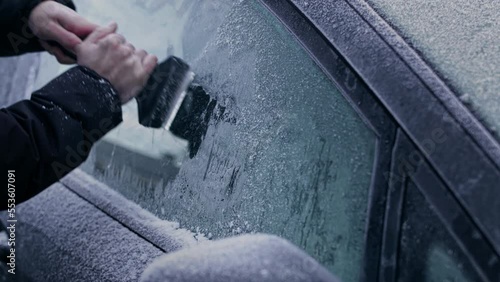 A man in a winter coat uses an ice scraper to remove heavy frost and ice from the windows and windshield of a frozen car on a sub-zero Winter morning.