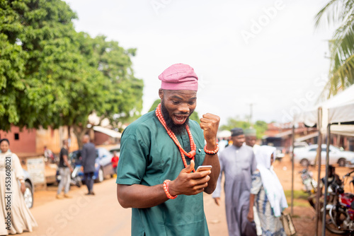 young African man excited looking at mobile at hand standing outside a rural street photo
