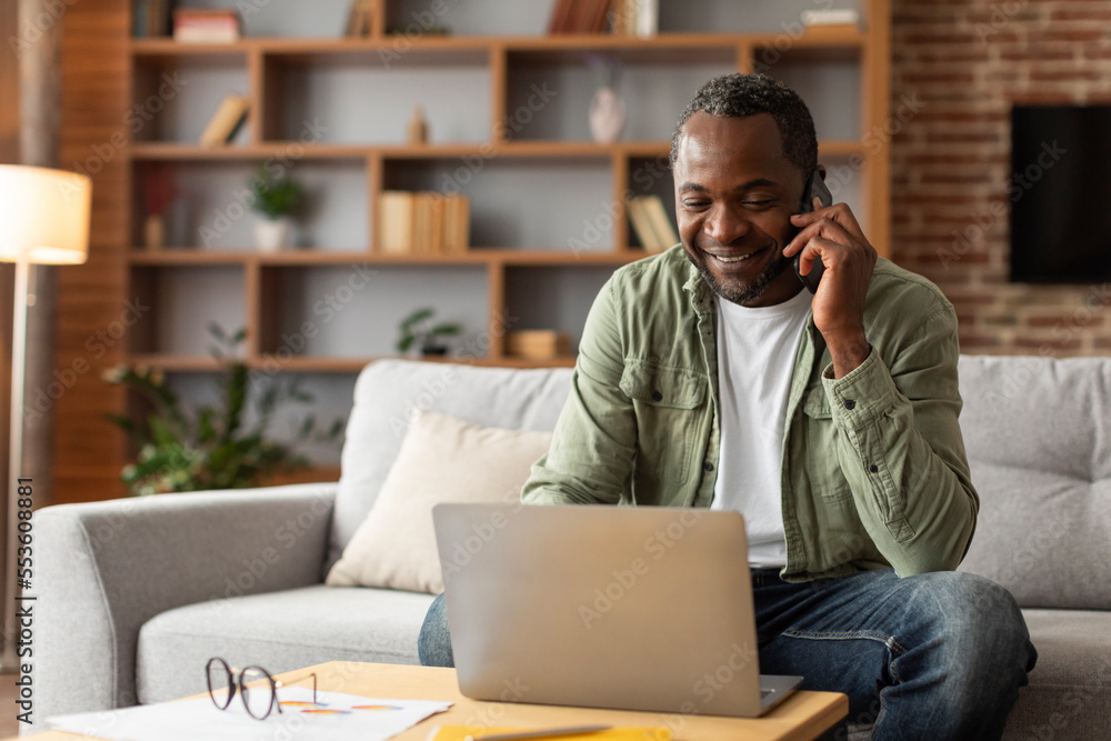 Smiling middle aged black male manager typing on computer, calling by phone, talk with client Smiling middle aged black male manager typing on computer, calling by phone, talk with client