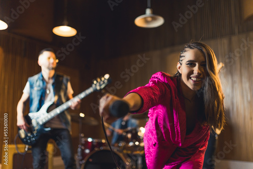 sing with us, young singer woman in pink clothes holding a microphone towards the camera and her band in the background. High quality photo