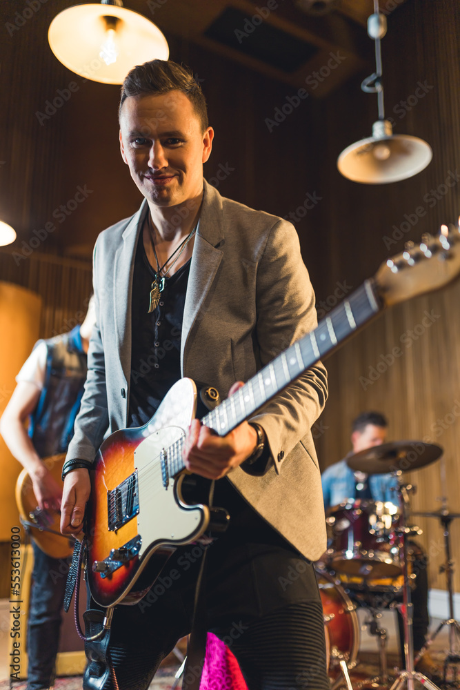 Fototapeta premium medium-full shot of a nice-looking young guitarist playing on repetition with his band, indoor music studio. High quality photo