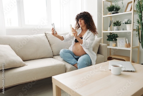 Pregnant woman smile blogger showing baby feeding bottle filming herself on phone, video chat sitting on sofa at home freelancer in last month of pregnancy
