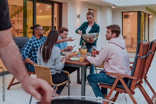 A group of young diverse people having dinner on the terrace of a modern house in the evening. Fun for friends and family. Celebration of holidays  weddings with barbecue.