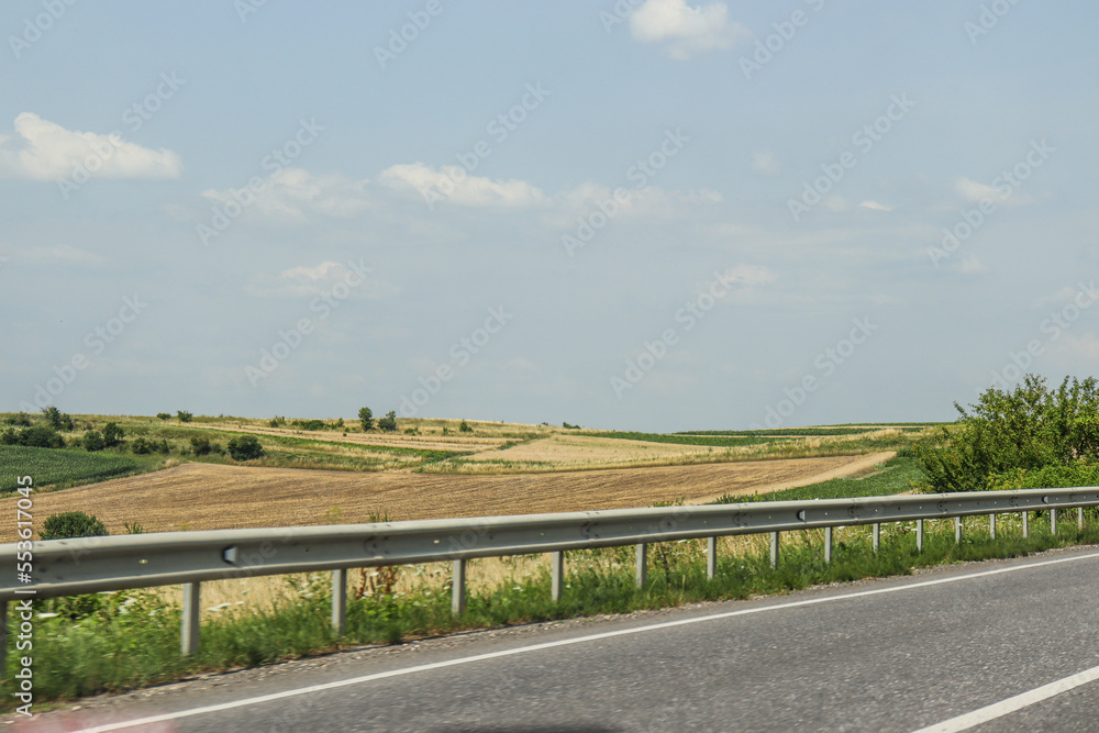 Yellow and green fields near the road, agriculture. Road fence. Greenery by road. Summer, travel, cars. Photo, country landscape, Ukraine