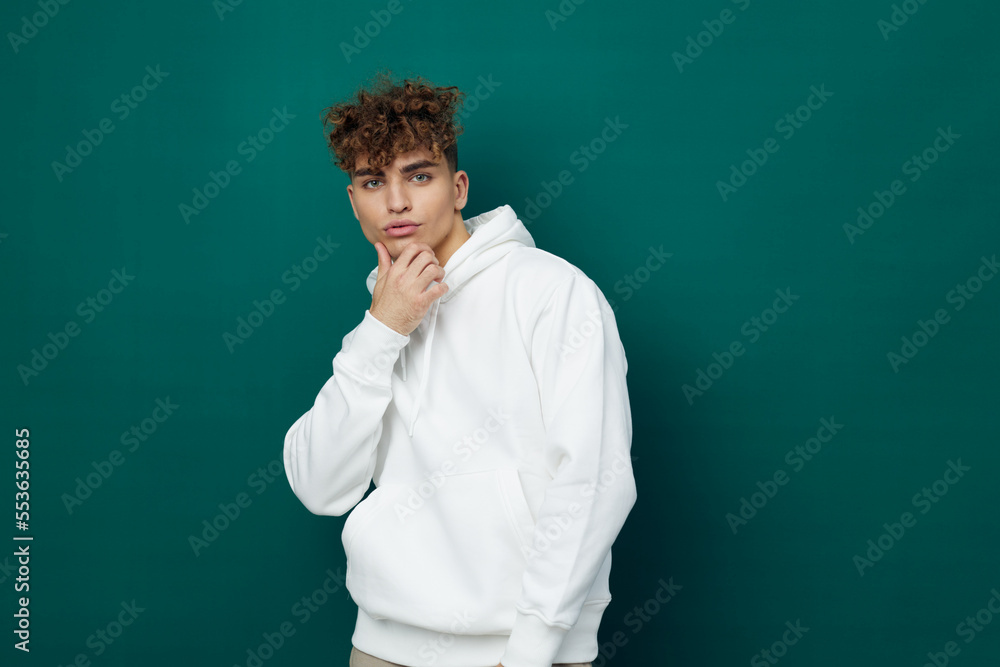 a handsome man stands on a green background in a white hoodie with a hood on his head and looks thoughtfully into the crowd, touching his chin with his hand a handsome man stands on a green background in a white hoodie with a hood on his head and looks thoughtfully into the crowd, touching his chin with his hand