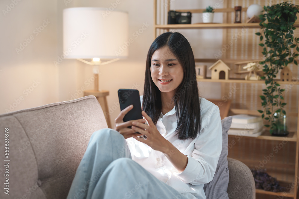 Beautiful Asian woman sitting on sofa in living room using mobile phone playing games and various entertainment programs at home holiday and having fun on vacation. Beautiful Asian woman sitting on sofa in living room using mobile phone playing games and various entertainment programs at home holiday and having fun on vacation.