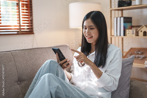 Beautiful Asian woman sitting on sofa in living room using mobile phone playing games and various entertainment programs at home holiday and having fun on vacation.