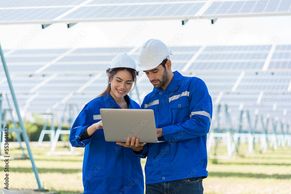Two male and female electrician worker holding laptop computer working in solar panels power farm between long rows array of photovoltaic solar cells panels. Green energy and Clean energy concept Two male and female electrician worker holding laptop computer working in solar panels power farm between long rows array of photovoltaic solar cells panels. Green energy and Clean energy concept
