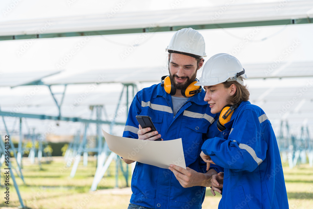 Two male and female electrician worker working in solar panels power farm between long rows array of photovoltaic solar cells panels. Green energy and Clean energy concept Two male and female electrician worker working in solar panels power farm between long rows array of photovoltaic solar cells panels. Green energy and Clean energy concept