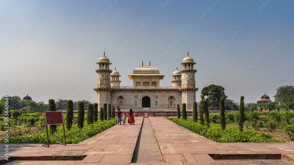 The  ancient mausoleum  of Itmad-Ud-Daula. The building is made of white marble with minarets. Arched openings, barred windows, ornaments, inlays of precious stones. People are walking along the path.
