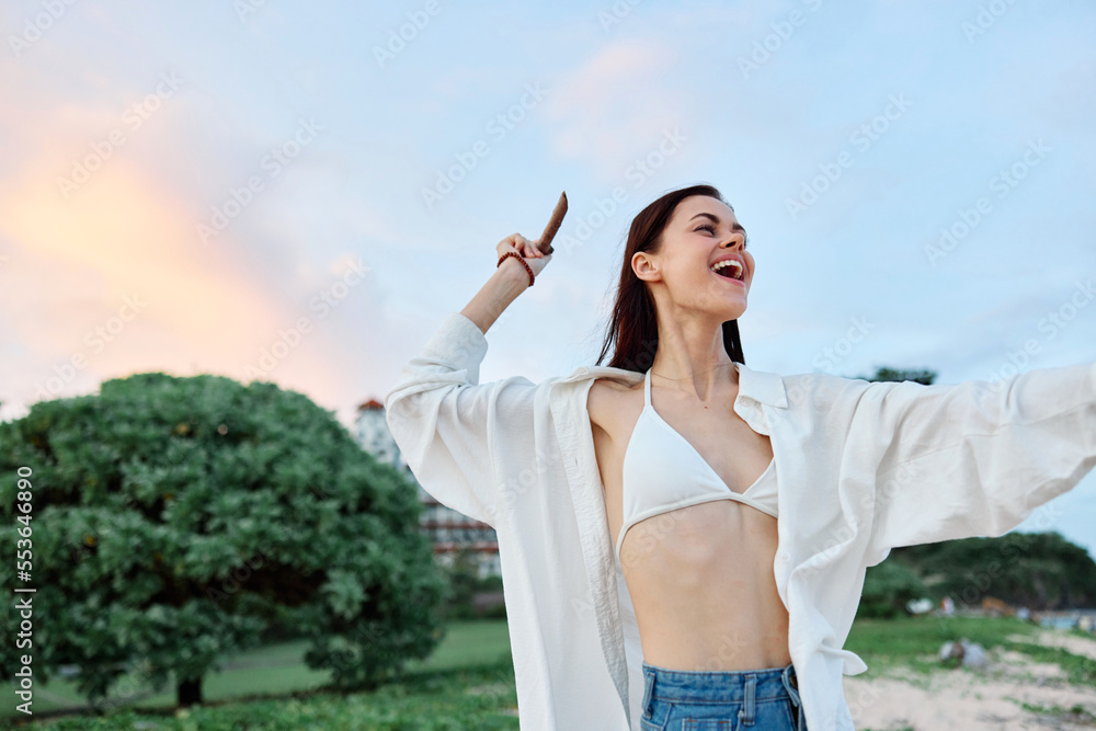 Brunette woman with long hair in a white shirt and jean shorts tan and happy fun throwing a stick smile with teeth against a backdrop of the beach and palm trees, vacation summer trip sunset sky