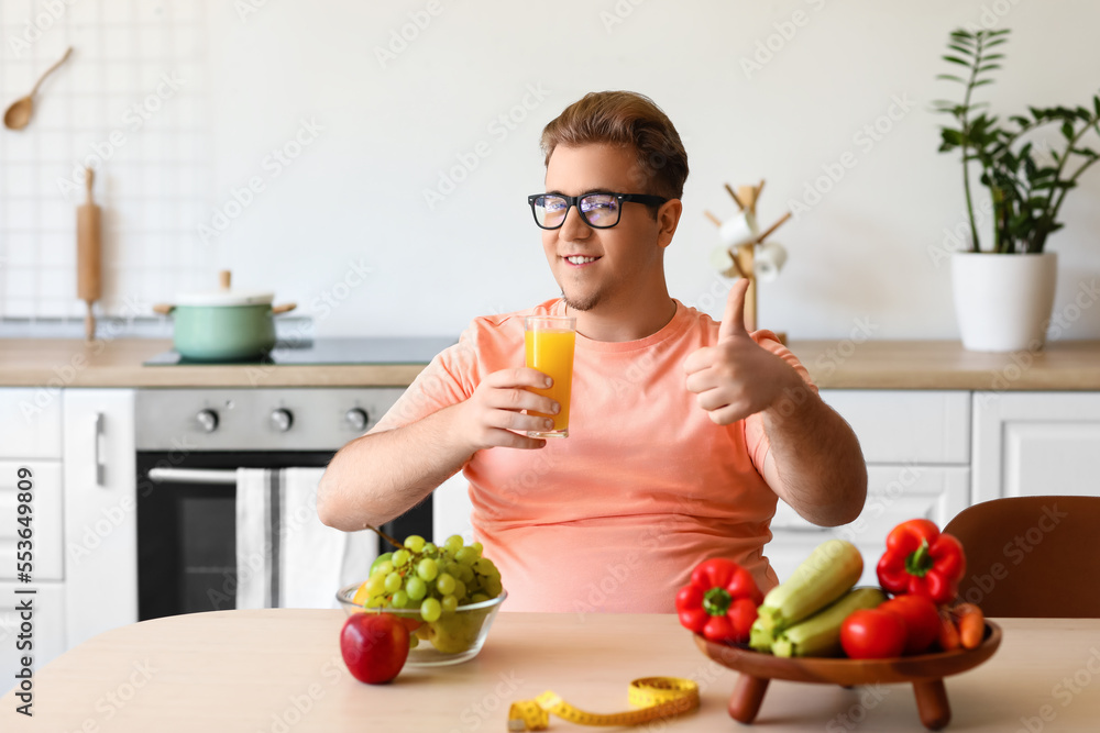 Young overweight man with glass of juice showing thumb-up in kitchen Young overweight man with glass of juice showing thumb-up in kitchen