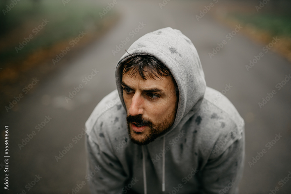 Young Caucasian athletic man pulling the hood over his head while having sports training in nature during the cold autumn day. Young Caucasian athletic man pulling the hood over his head while having sports training in nature during the cold autumn day.