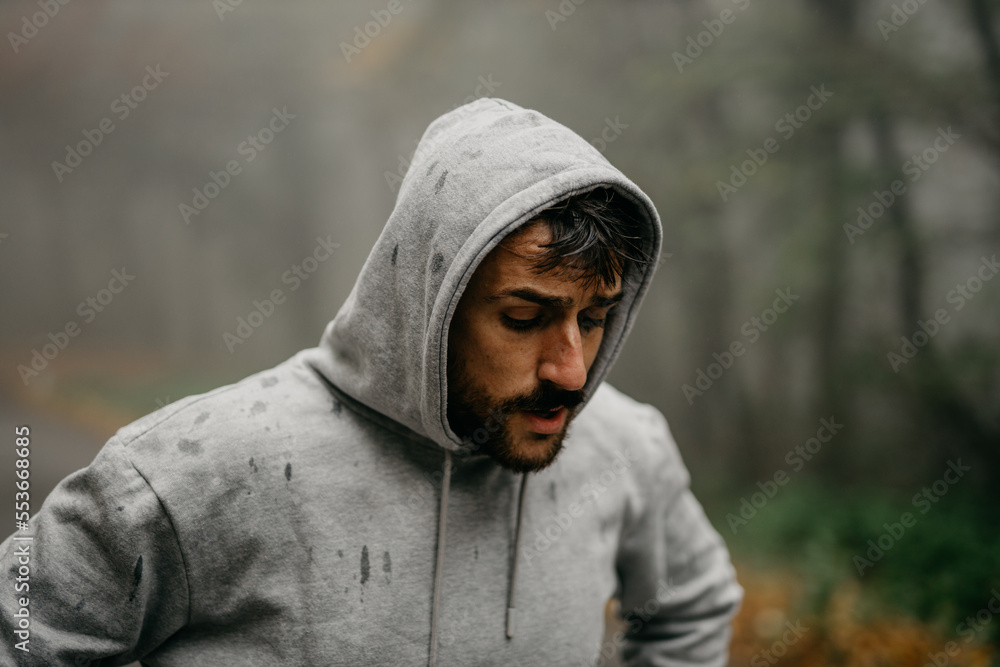 Young Caucasian athletic man pulling the hood over his head while having sports training in nature during the cold autumn day. Young Caucasian athletic man pulling the hood over his head while having sports training in nature during the cold autumn day.