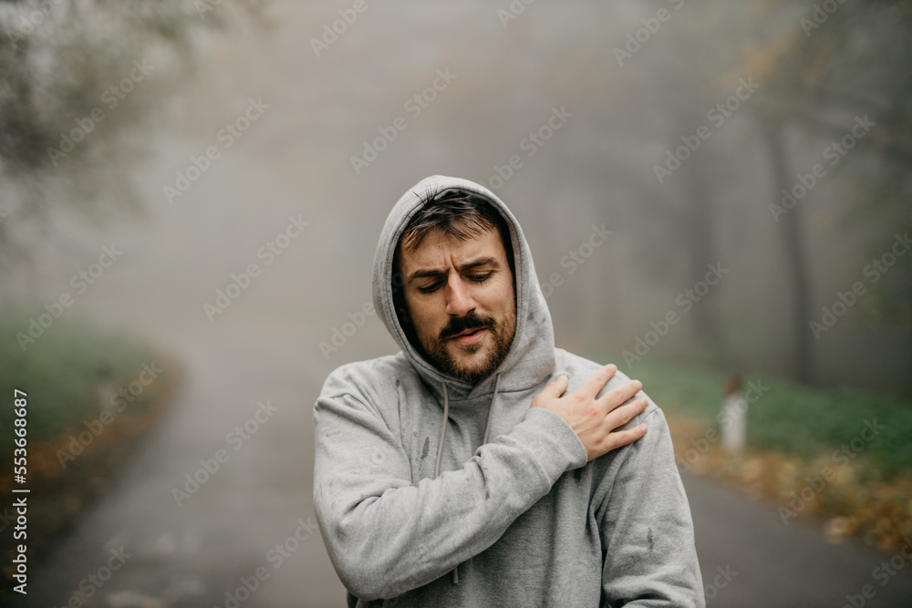 Young Caucasian athletic man pulling the hood over his head while having sports training in nature during the cold autumn day. An athlete is feeling pain in the shoulder Young Caucasian athletic man pulling the hood over his head while having sports training in nature during the cold autumn day. An athlete is feeling pain in the shoulder