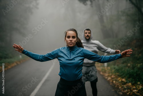 Two people in sports outfits have an active HIIT workout in the forest. Female in front, and a man in the background, both doing jumping jacks to warm up muscles.