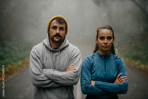 Portrait of a man and woman standing and looking at the camera with arms crossed before/after jogging in the forest.