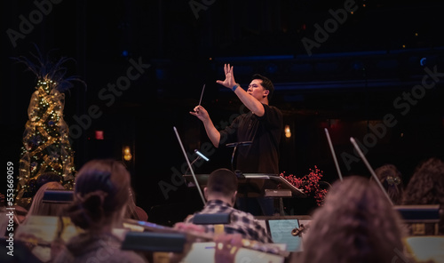 Middle-aged male conductor directing rehearsal of Christmas concert; silhouettes of musicians in foreground; Christmas background