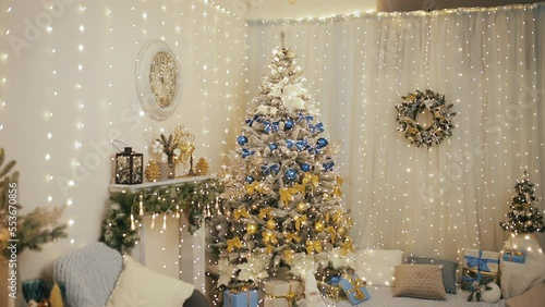 Christmas, New Year 2023 in Ukraine. Decorated fir-tree with blue bulbs and golden yellow bows. Fireplace, golden reindeer toys, clock and wreath on wall