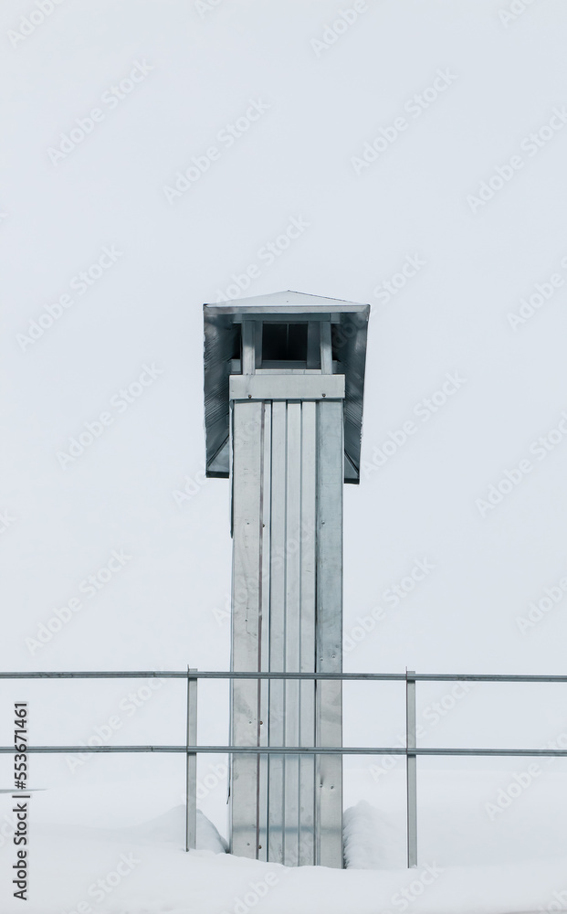 Smoke, rectangular exhaust pipe on the roof of a modern house. The roof of the building is covered with white snow against the gray sky. Cloudy, cold winter day, soft light.