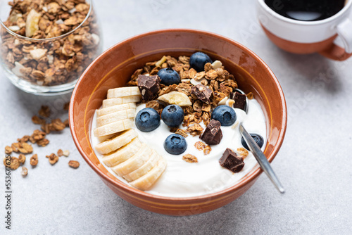 Yogurt with granola, banana, blueberries and dark chocolate in bowl, grey cocnrete table background, closeup view. Healthy food photo