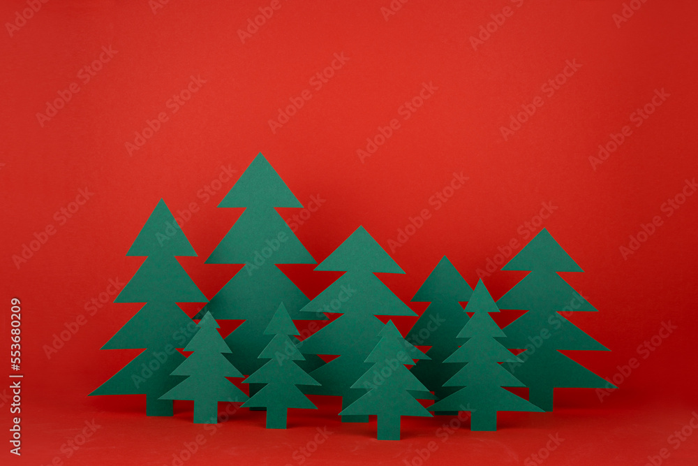 Obraz premium Traditional Christmas festive background with cartoon paper spruces as forest on red backdrop in elegant baby style. New Year mockup for advertising, design, card, poster, flyer.