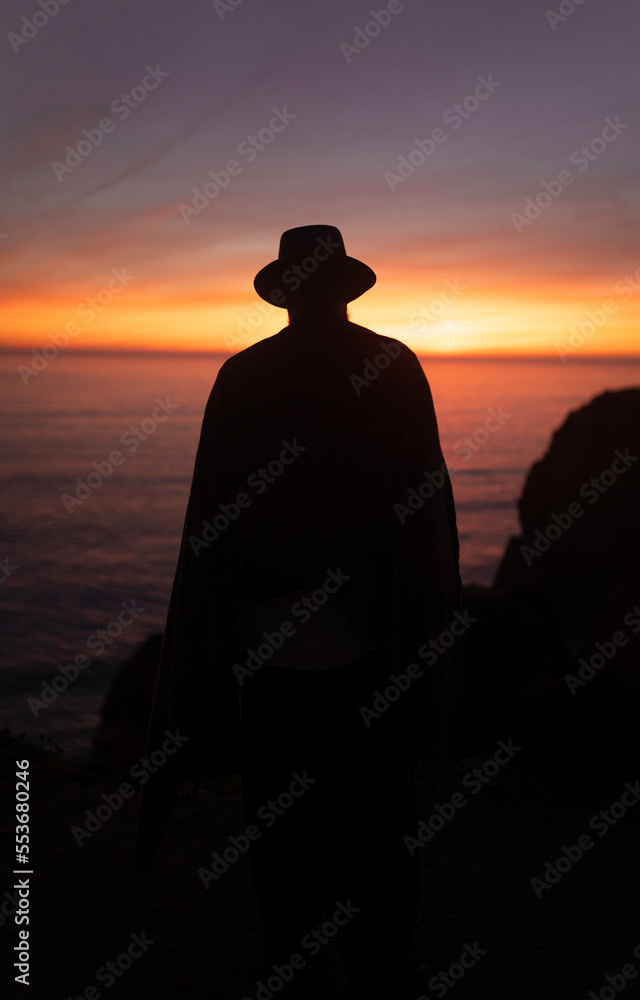 Silhouette of a man watching the ocean during the sunset wearing a poncho and a hat Silhouette of a man watching the ocean during the sunset wearing a poncho and a hat